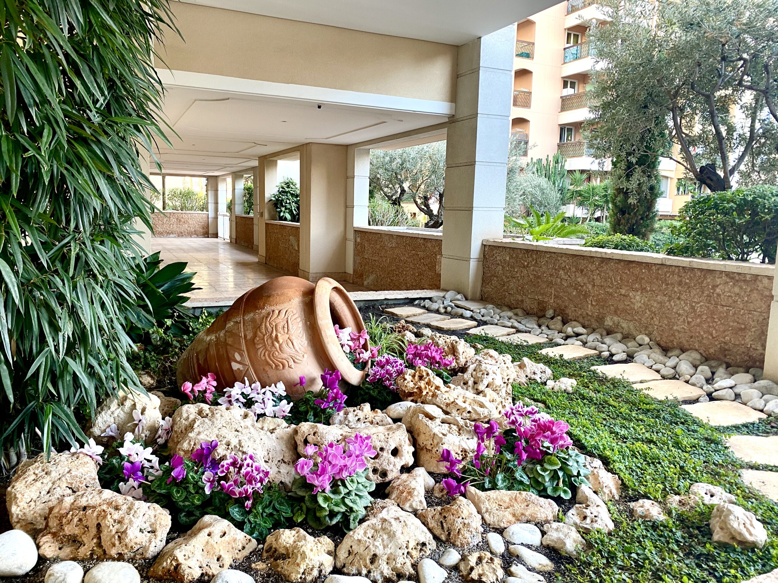 Garden area of Terrasses du Port building. Green plants and pink flowers set up around a path made of tiles