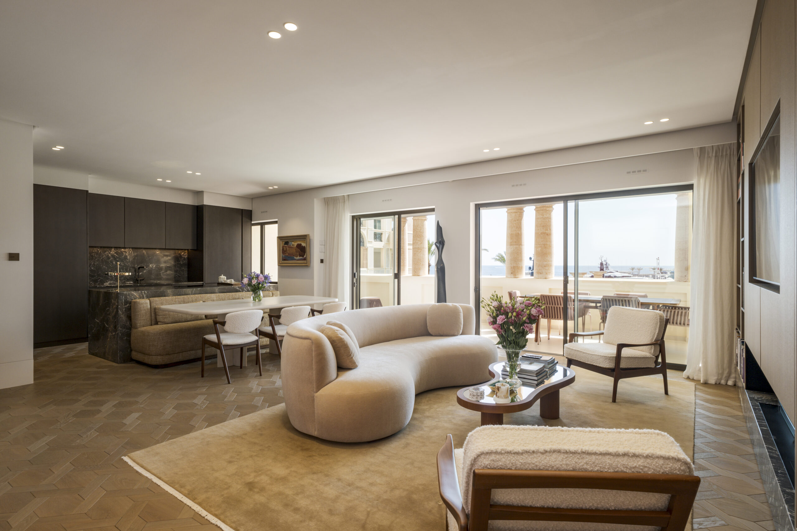 Bright open plan living room in a Seaside Plaza apartment in Monaco with curved cream sofa, marble kitchen island and sliding doors opening onto a terrace with sea views
